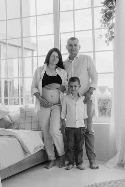 Samimi bir aile fotoğrafı, hassas bir beklenti anını yakalıyor. Siyah bluz ve düğmesiz beyaz gömlek giyen hamile anne elini nazikçe karnına koyar. Beyaz gömlekli baba, onun arkasında korunuyor.