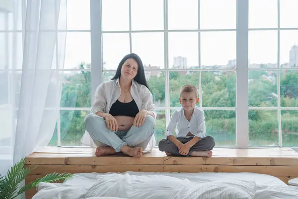 Samimi bir aile fotoğrafı, hassas bir beklenti anını yakalıyor. Siyah bluz ve düğmesiz beyaz gömlek giyen hamile anne elini nazikçe karnına koyar. Beyaz gömlekli baba, onun arkasında korunuyor.