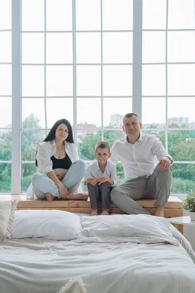 Samimi bir aile fotoğrafı, hassas bir beklenti anını yakalıyor. Siyah bluz ve düğmesiz beyaz gömlek giyen hamile anne elini nazikçe karnına koyar. Beyaz gömlekli baba, onun arkasında korunuyor.