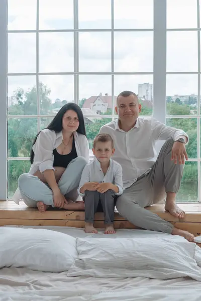 Samimi bir aile fotoğrafı, hassas bir beklenti anını yakalıyor. Siyah bluz ve düğmesiz beyaz gömlek giyen hamile anne elini nazikçe karnına koyar. Beyaz gömlekli baba, onun arkasında korunuyor.