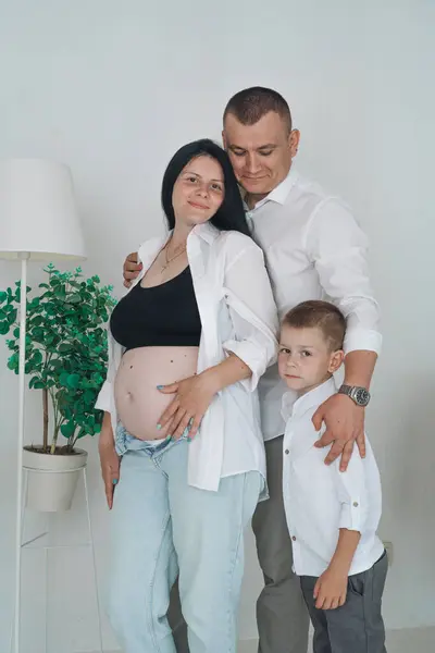 Samimi bir aile fotoğrafı, hassas bir beklenti anını yakalıyor. Siyah bluz ve düğmesiz beyaz gömlek giyen hamile anne elini nazikçe karnına koyar. Beyaz gömlekli baba, onun arkasında korunuyor.