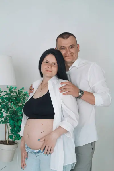 Samimi bir aile fotoğrafı, hassas bir beklenti anını yakalıyor. Siyah bluz ve düğmesiz beyaz gömlek giyen hamile anne elini nazikçe karnına koyar. Beyaz gömlekli baba, onun arkasında korunuyor.