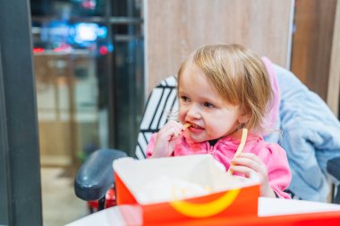 A funny little girl is enjoying fast food, eating fries and a hamburger, and drinking juice. Her playful grimace adds an extra touch of humor to the scene. clipart