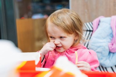 A funny little girl is enjoying fast food, eating fries and a hamburger, and drinking juice. Her playful grimace adds an extra touch of humor to the scene. clipart