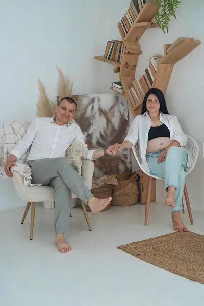 Samimi bir aile fotoğrafı, hassas bir beklenti anını yakalıyor. Siyah bluz ve düğmesiz beyaz gömlek giyen hamile anne elini nazikçe karnına koyar. Beyaz gömlekli baba, onun arkasında korunuyor.