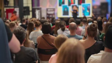 Bir seyirci eflatun bir odada toplanmış sahnede bir sunum izliyor. Bu etkinlik, büyük bir kalabalığın ekran cihazını paylaştığı bir eğlence armaşıdır.