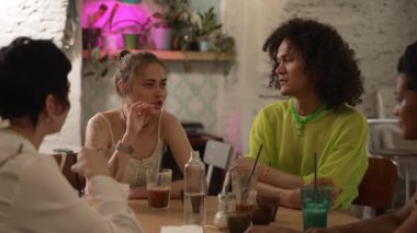 4K Güzel Igbt insanları modern kafedeki masada oturup konuşuyorlar. Çekici genç kadınlar, gülümseyen, konuşan, kutlama yapan ve iç mekanda masa başında oturan bir erkek. Neşeli gay arkadaşlar takılır