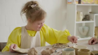 Down sendromlu bir çocuk kil atölyesine katılıyor ve sanatına odaklanıyor. Şu an sanat eğitiminin ve gelişim desteğinin öneminin altını çiziyor. Konsept: gelişimsel büyüme, sanat becerileri
