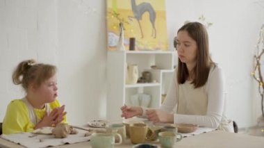 Down sendromlu çocuk ve eğitmen ışık dolu stüdyoda kil parçaları yaratıyor. İlk sanat eğitiminin ve interaktif öğrenmenin faydalarını ön plana çıkarıyor. Yaratıcı beceri geliştirme kavramı