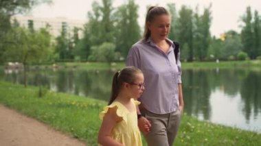 Down sendromlu anne ve kızı göl boyunca el ele yürüyorlar. Annesi açık mor gömlek giyerken, kız sarı elbise ve kırmızı gözlük takıyor. Sahne özel ihtiyaç duyulan çocuklar kavramını yakalar