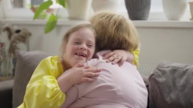 Down sendromlu gülen çocuk sıcak oturma odasında annesine sarılıyor. Sahne, kabul edilmeyi ve aile içinde sosyal katılımın önemini yansıtır. Kavram: Toplumsal kabul, kapsama, destek