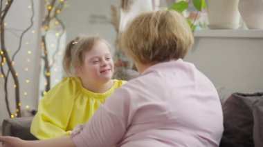 Down sendromlu çocuk ve anne sıcak yuva ortamında kucaklaşırlar. Görüntü, aşk, kabullenme ve sosyal içerikleri yansıtır. Kavram: Kabullenme, aidiyet, sosyal bağlantı. Yüksek