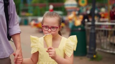 Down sendromlu sarı elbiseli genç bir kız annesiyle lunaparkta otururken dondurma yemekten hoşlanıyor. Sahne, çocuğu özel ihtiyaçlarla büyütmenin basit zevklerini yakalar. Yüksek kalite