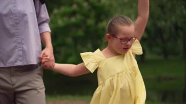 Down sendromlu, sarı elbiseli, annesinin elini tutan ve sevinçle kolunu kaldıran genç bir kız. Parktaki göl manzarası, özel çocuk yetiştirme kavramını yansıtıyor. Yüksek