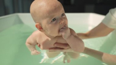 Profesyonel yüzme hocası, gülümseyen bebeğe modern küvette yardım ediyor. Güneş ışığı, parlak ayarı güçlendiriyor. Bebek çiğniyor, eğitmen destekleyici rehberlik sunuyor, güvenliği ve yüzme becerilerini vurguluyor