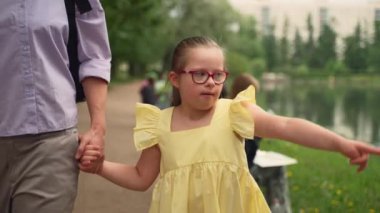 Down sendromlu, sarı elbiseli genç bir kız annesiyle parkta yürüyor. Annesinin elini tutarken kolunu heyecanla sallıyor. Kapsama ve kabullenme kavramları. Yüksek kalite