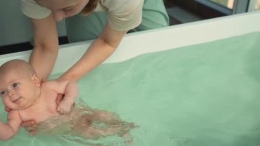 Üç aylık küçük bir kızla erken gelişim yüzme seansı, eğitmenlerin rehberliği apaçık ortada. Bebek arkada yüzüyor, şehir penceresinden bakıyor. Sahne temel büyümeyi ve bakımı vurguluyor