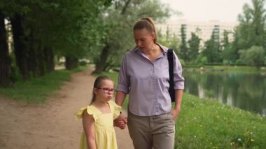 Down sendromlu, gözlüklü ve sarı elbiseli genç bir kız annesiyle el ele tutuşarak parkta yürüyor. Yeşilliklerle çevrili kız yürürken kolunu kaldırıyor. Kapsama kavramları, kabullenme