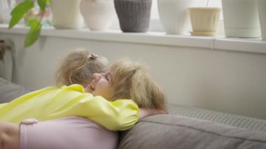 Down sendromlu mutlu bir kız, gülen annesine oturma odasında sarılıyor. Sahne, aile sevgisi ve bağlılık yoluyla duygusal refahı ön plana çıkarır. Konsept: Ruh sağlığı, destek, duygusal güvenlik