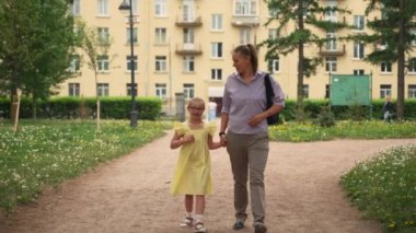 Down sendromlu anne ve kızı parkta el ele yürüyorlar. Anne mor bluz giyerken kız sarı elbise ve kırmızı gözlük takıyor. Arkaplanda bina ve yeşillik