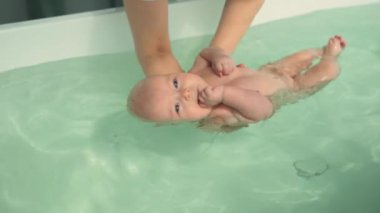 Anne, üç aylık bebeğini nazikçe yıkıyor ve rahat bir şekilde yüzmesini sağlıyor. Parlak doğal ışık odayı dolduruyor, annelik sevgisini ve barışçıl yakınlaşma anını vurguluyor. Yüksek kalite 4k