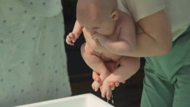 Anne, elinde havluyla üç aylık bir bebeği banyodan sonra eğitmenlikten alıyor. Eğitmen, bebeği nazikçe sudan kaldırıyor. Bakım, güvenlik ve banyodan anneye geçiş konularını vurguluyor.