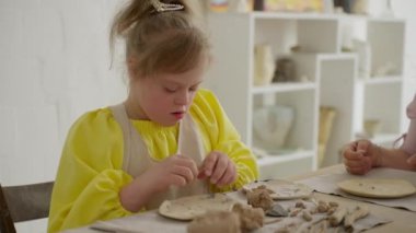 Down sendromlu bir çocuk, annesinin rehberliğinde, sanat stüdyosunda kil şekillendiriyor. Şu an sanat ortamında erken eğitim faaliyetlerinin önemini gösteriyor. Konsept: erken eğitim, yaratıcı gelişme