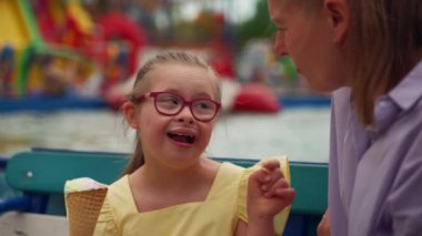 Down sendromlu gülümseyen çocuk annesiyle konuşurken dondurmanın tadını çıkarıyor. Suya yakın otururlar, canlı çekimlerle çevrilidirler. Yüksek kalite 4k görüntü