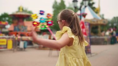 Sarı elbiseli, Down sendromlu bir çocuk lunaparkta yürürken fırıldak sallıyor. Sahne enerjiyi, eğlenceyi ve canlı atmosferi vurgular. Yüksek kalite 4k görüntü