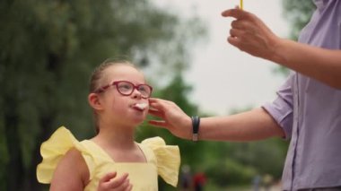Sarı elbiseli Down sendromlu bir kız parkta mor gömlekli bir kadının yanında dururken pamuk şeker yemekten hoşlanıyor. Sahne basit zevkleri, kahkahaları ve bağlantıları vurgular. 4k görüntü