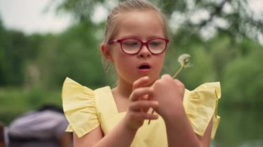 Down sendromlu pembe gözlüklü ve sarı elbiseli bir kız göl kenarında yeşil alanda karahindiba yiyor. Sahne masumiyeti ve duygusal refahı yansıtır. Yüksek kalite 4k görüntü