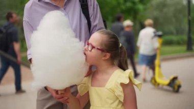 Down sendromlu pembe gözlüklü ve sarı elbiseli bir kız parkta yürürken mor gömlekli bir kadın tarafından tutulan pamuk şekere uzanır. Kaygısız bir an. Yüksek kalite 4k görüntü