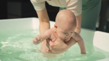 Günışığı ve modern küvette yüzme dersinde eğitmen mutlu bebeğe rehberlik eder. Bebek gülümsüyor ve elinde çiğniyor, eğitmen güvenli, destekleyici bir ortam sağlıyor, etkin bir öğretim sağlıyor. Yüksek kalite
