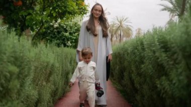 Anne ve oğlu, aile arazisinin yemyeşil bahçesinde birlikte yürüyorlar. Kadın ipek eşarp taşırken çocuk oyuncak araba tutuyor. Aileyi ön plana çıkaran sakin ve samimi bir sahne yaratıyor. Yüksek kalite 4k görüntü