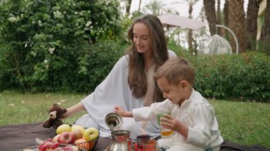 Anne ve oğlu yemyeşil Fas bahçesinde birlikte oturup piknik battaniyesinde yiyecek ve içecekleri keşfediyorlar. Canlı yeşilliklerle çevrili meraklı ve eğlenceli anları paylaşıyorlar. Yüksek kalite 4k görüntü