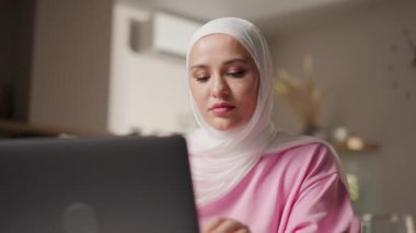 Beyaz tesettürlü Müslüman bir kadın dizüstü bilgisayarla oturmuş, düşünceli bir şekilde video içeriğini izliyor. Odaklanmış görünüyor, muhtemelen webinar ya da özel derse katılıyor. Büyüme ve dijital eğitim kavramı. Yüksek