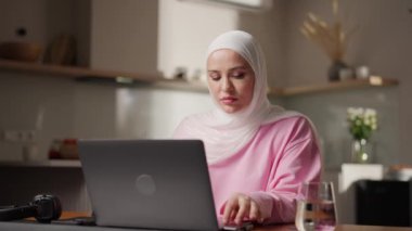 Mutfakta oturan beyaz tesettürlü Müslüman kadın internette dizüstü bilgisayarını karıştırıyor. Rahat duruşu internette boş zamanlarının tadını çıkardığını gösteriyor. Günlük dijital yaşam tarzı ve ev konforu kavramı. Yüksek