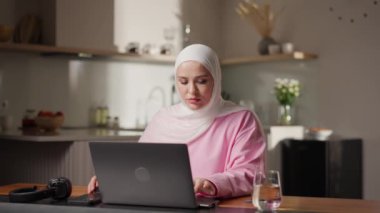 Beyaz tesettürlü Müslüman kadın rahat mutfaktaki dizüstü bilgisayardan dikkatlice okuyor. Düşünceli ifadesi internetten öğrenmeyi ya da araştırmayı yansıtıyor. Evden eğitim ve bilgi edinme kavramı