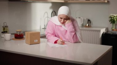Kamera, Müslüman kadın portresinden, mutfak tezgahında oturan pembe tesettürlü kadının dönüş formu üzerindeki çalışmalarını daha geniş açıdan görebilmek için hareket ediyor. Odaklanmış ve sakin görünüyor. Çevrimiçi alışveriş ve geri dönüşler kavramı