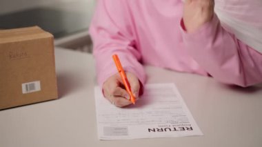 Dönüş formu, pembe tesettürlü Müslüman kadın turuncu kalemle doldururken yakın planda gösteriliyor. Onun eli kağıt üzerinde sürekli hareket eder. Dikkatli çevrimiçi dönüş süreci ve e-ticaret kavramı. Yüksek kalite