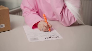 İade formunu beyaz masaya yakın plan çek. Pembe tesettürlü Müslüman kadın turuncu kalemle detayları yazıyor. Belge ve el yazısı net bir şekilde odaklanmış durumda. Hassasiyet ve belgelendirme kavramı. Yüksek kalite
