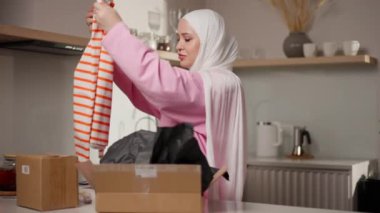 Pembe tesettürlü Müslüman kadın, rahat mutfağındaki teslimat kutusundan çizgili uzun kollu gömleği inceliyor. Detayları kontrol edip kaliteyi sağlıyor. Ürün denetimi ve müşteri deneyimi kavramı