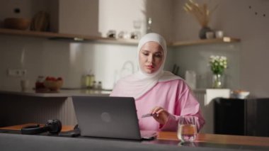 Pembe tesettürdeki Müslüman kadın mutfaktaki rahat tesettürde, elinde banka kartı tutarken dizüstü bilgisayarına detaylar giriyor. Satın almayı güvenli bir şekilde tamamlamaya odaklanıyor. Dijital işlemler ve e-ticaret kavramı