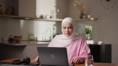 Mutfak masasında dizüstü bilgisayarında beyaz tesettürlü Müslüman bir kadın. Odaklanmış görünüyor, ekrandaki bilgiyi analiz ediyor. Profesyonel serbest çalışma kavramı ve ev ortamında yoğunlaşma. Yüksek
