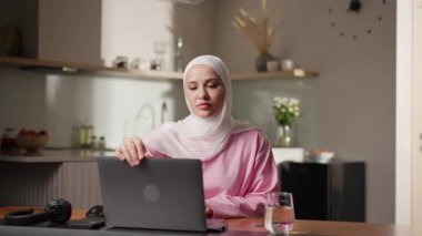 Beyaz tesettürlü Müslüman kadın, rahat mutfağın ahşap masasında otururken dizüstü bilgisayarı açıyor. Verimli bir seansa hazırlanıyor, çalışmaya ya da çalışmaya hazır. Uzaktan çalışma kavramı ve evde çevrimiçi öğrenim