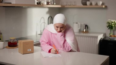 Kamera geniş bir mutfak manzarasından, dönüş formunu dolduran pembe tesettürlü Müslüman kadının yakın çekimine zum yapıyor. O, hassasiyet ve konsantrasyonla yazar. E-ticaret ve geri dönüş süreci kavramı. Yüksek