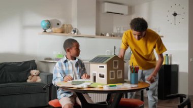 Küçük kardeş güneş enerjisi modeli üzerinde çalışmaya devam ederken ağabeyi yaklaşmakta, gözlemlemekte ve ilerlemeyi onaylamaktadır. Akıl hocalığı, takım çalışması ve yenilenebilir enerji eğitimi kavramı. Yüksek kalite 4k
