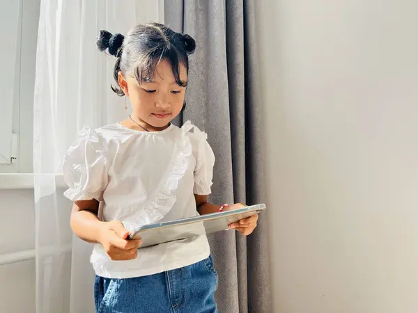 stock image Asian little girl standing near the window holding mobile tablet. Screen time, phone addictive concept