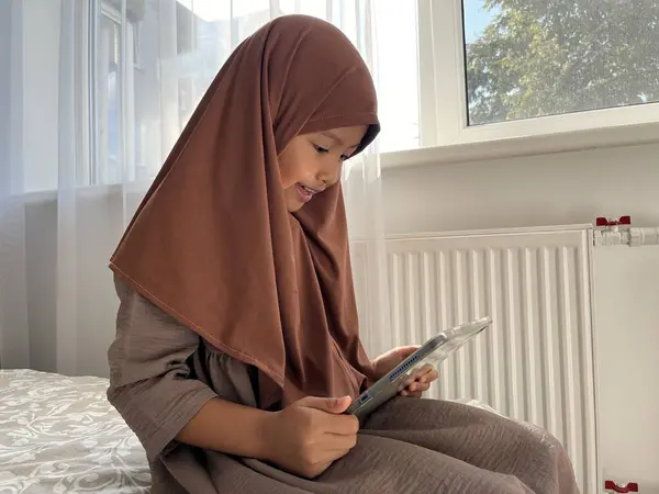 Asyalı Müslüman küçük kız tesettüre binmiş pencerenin kenarında oturmuş tablette film seyrediyor. 