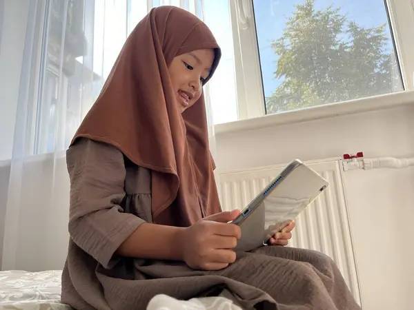 Asyalı Müslüman küçük kız tesettüre binmiş pencerenin kenarında oturmuş tablette film seyrediyor. 